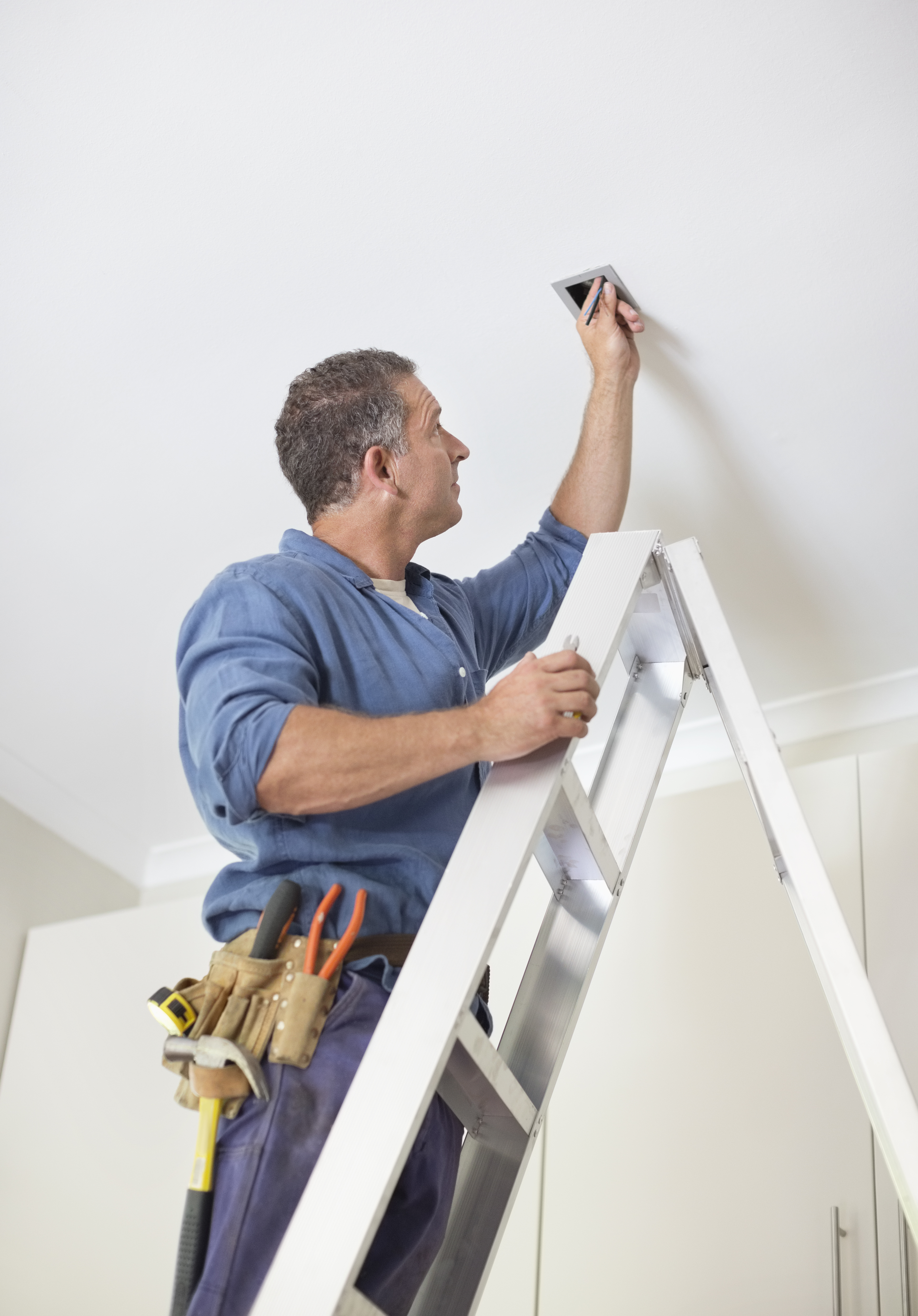 A skilled handyman is seen perched atop a ladder, meticulously working on installing a lighting fixture, demonstrating expertise and attention to detail in home